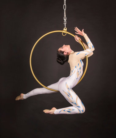 A slender,dark-haired girl - an air acrobat in a white and blue suit, performs exercises in an air ring. Studio shooting on a dark background.の写真素材