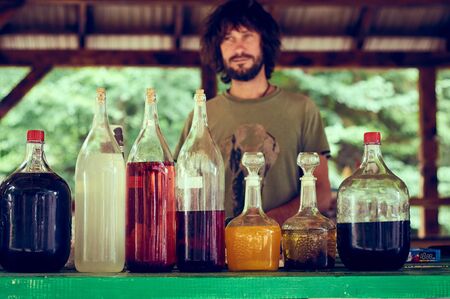Karpaty, Ukraine, Shipot waterfall - July 20, 2019. A colorful Ukrainian man with a beard sells home-made alcohol outdoors near Shipot waterfall. Beautiful glass bottles of different shapes and sizes.のeditorial素材