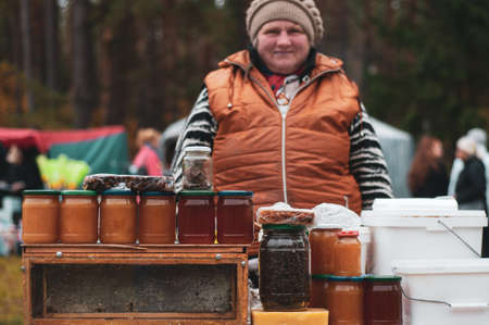 Belarus, urban village Glusha - October 5, 2019. An elderly woman sells natural honey in jars at the autumn harvest festival.のeditorial素材