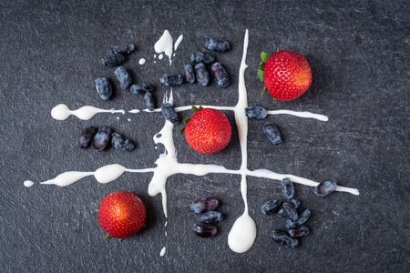 Red strawberries and dark blue honeysuckle on a dark stone board with white sour cream. Game of tic tac toe with fresh berries.の写真素材