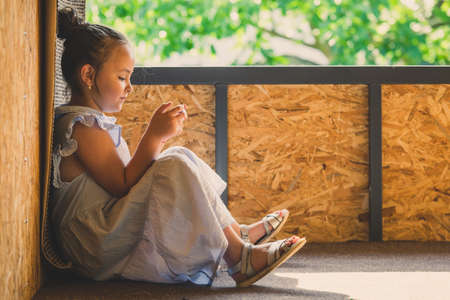 Child smiling girl on a summer afternoon on the porch playing with her smartphone. Reads or sends...
