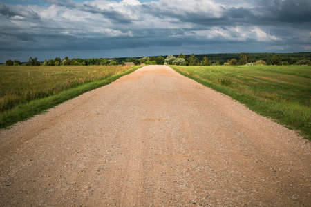A strip of country sandy road through the field to the village. Sun through the clouds. Belarusian summer landscape.の写真素材