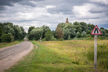 A strip of country road through the field to the trees behind which you can see the dome of the old ruined church. Belarusian summer landscape with a dramatic cloudy sky.の写真素材