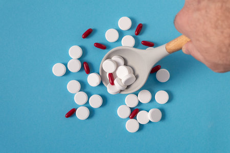 A hand with a spoon mixes white vitamin pills and red capsules on a blue background with space to insert text. medicine and health.の写真素材