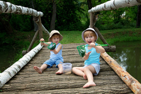 Adorable Little Twin Brothers Sitting on a Wooden Bridge and Holding a Fishnet Full of Fish at the Lake Facing Cameraの写真素材