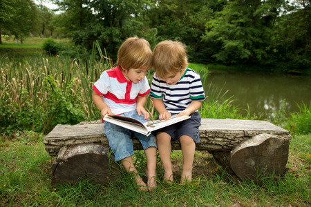 Adorable Little Twin Brothers Sitting on a Wooden Bench and Reading a Book Very Carefully  Near the Beautiful Lake at Summerの写真素材