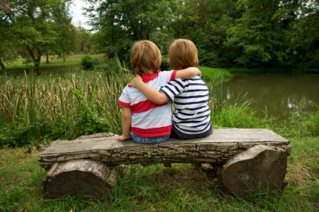 Adorable Little Twin Brothers Sitting on a Wooden Bench, Embracing Each Other and Looking at Beautiful Lake at Summerの写真素材