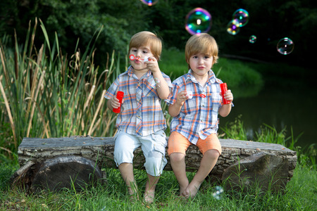 Little Twin Brothers Sitting on Wooden Bench and Blowing Soap Bubbles in Summer Park の写真素材