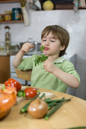 Adorable Little Boy Making Funny Face While Eating Parsley in the Kitchen at Homeの写真素材