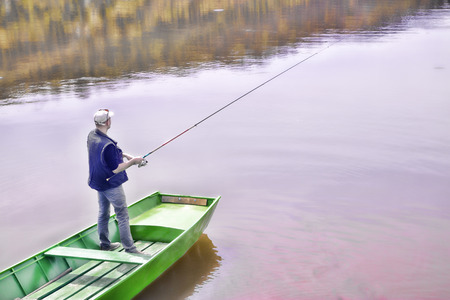 Fisherman Casting A Fishing Rod From The Green Boat On The Lake And Patiently Waiting For Fish To Take A Bait, Water Reflectionの写真素材