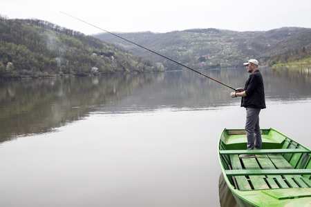 Side View Of Sport Fisher With Cap And Vest Fishing From Green Canoe And Enjoying His Vacation On Beautiful Lakeの写真素材