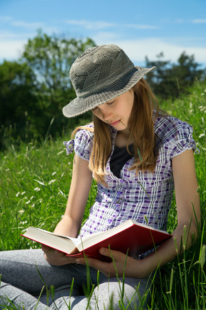 Smart Teenage Girl Sitting In The Grass With Legs Crossed Reading A Book Outdoors On Sunny Day Closeupの写真素材