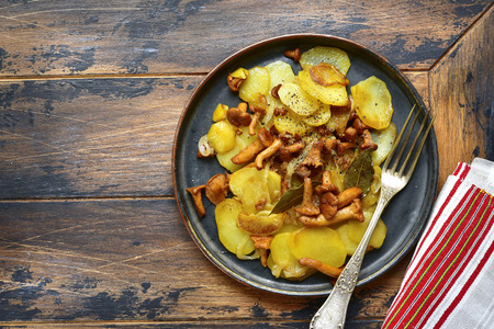 Potato slices fried with chanterelle mushrooms and onion on a black plate over rustic wooden background.Top view.の写真素材