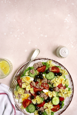 Pasta salad with tuna, cherry tomatoes, cucumber, black olives and boiled eggs on a plate over light pink slate, stone or concrete background.Top view with copy space.の写真素材