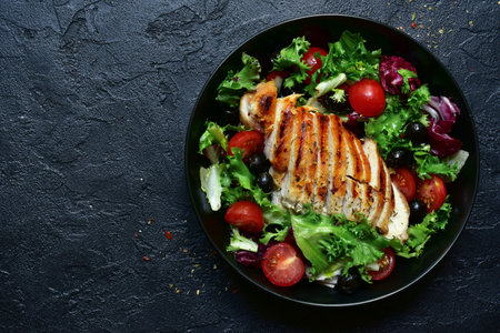 Grilled chicken fillet with vegetable salad in a black bowl on a dark slate, stone or concrete...