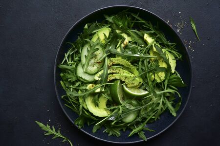 Green vegetable salad with arugula, cucumber and avocado in a black bowl on a dark slate, stone or concrete background. Top view with copy space.の写真素材