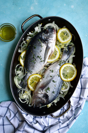 Raw fresh dorado prepared for baking with onion, lemon and thyme in a skillet pan. Top view with...