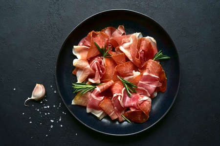 Slices of prosciutto di parma or jamon serrano (iberico) on a black plate on a dark slate, stone or concrete background. Top view with copy space.の写真素材