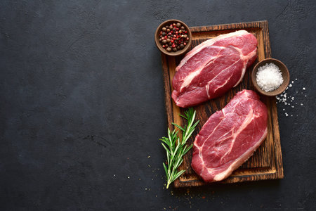 Raw organic marbled beef steaks with spices on a wooden cutting board on a black slate, stone or...