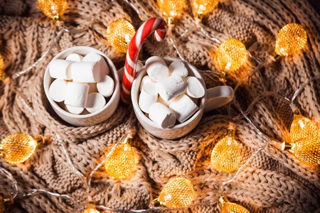Christmas background with cocoa or coffee with marshmallows and candy in a white cup on a brown knitted winter scarf and a glowing golden garland. Beautiful concept of home comfort and warmthの写真素材