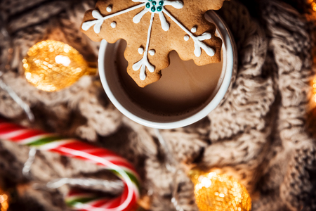 Christmas background coffee in a white cup and gingerbread snowflakes with candy on a brown knitted winter scarf and a glowing golden garland. Beautiful home Holiday drinkの写真素材