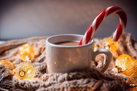 Christmas background coffee in a white cup with candy on a brown knitted winter scarf and a glowing golden garland. Beautiful home Holiday drinkの写真素材