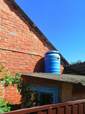 blue barrel. on the roof against the blue sky and red brick. sunny day. improvement of a private house. container made of plastic for heating water, outdoor showerの写真素材