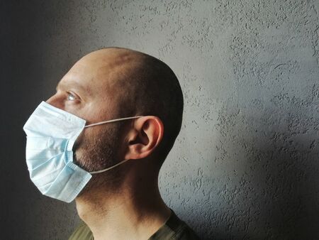 a masked man in profile looks up. covid-19. young man in a protective medical mask in isolation - sideways on a gray background. pandemic, coronavirus - place for textの写真素材