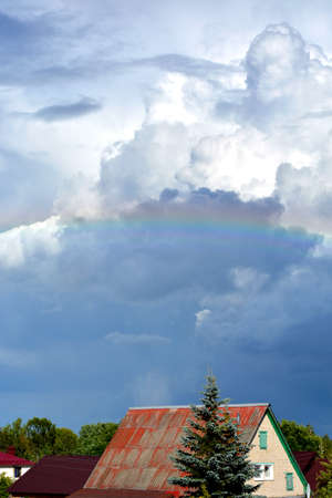 Rainbow over the roofs of houses in the blue sky. White clouds float across the sky. Picturesque landscape.の写真素材