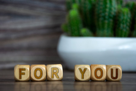 The word FOR YOU on wooden cubes on a blurred background near a flower in a pot. Defocusing, concept. Alphabet letter blocks on wooden reflective background.の写真素材