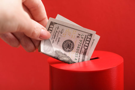 Female hand puts dollar into slot of red donation box. Concept of donorship, life saving or charity. Concept of sincere devotion to faith.の写真素材