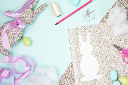 Easter flat lay. DIY. Soft decorative toy Easter Bunny made of cotton fabric in small flower, decorated with pink satin ribbon bow. Supplies needed to make craft. Layout on blue background. top viewの写真素材