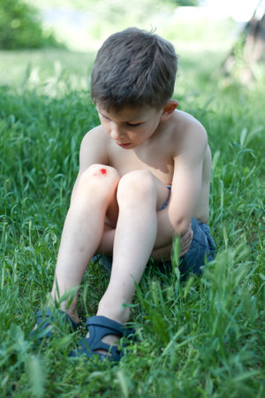 Portrait of preschool boy sitting on grass with fresh bleeding wound on knee. child fell, scratched skin on knee, hurt, upset. Children injuries in summer outdoors.の写真素材