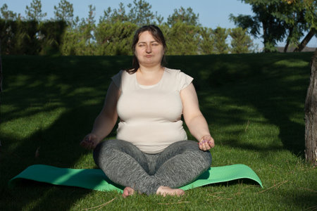 Portrait of overweight Caucasian middle-aged woman practicing yoga in nature. Fat woman meditating, relaxing in lotus position, sitting on fitness mat. Calm, relaxed, zen, stop stress, here and nowの写真素材