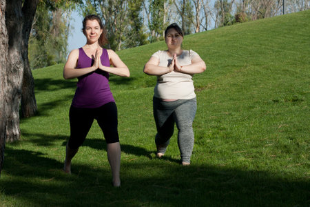 Two middle-aged women practice yoga in city park in warrior pose, standing barefoot on grass. Namaste. Healthy lifestyle, fitness, Pilates, weight loss. overweight woman doing yoga with yoga trainer.の写真素材