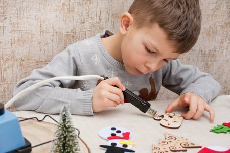 Merry Christmas and Happy New Year. preschool boy burning ornament on wooden blank using pyrograph, sitting at table at home. Child preparing decorations for Christmas tree. Children's creativity.の写真素材