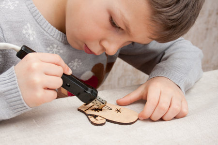 Merry Christmas and Happy New Year. preschool boy burning ornament on wooden blank using pyrograph, sitting at table at home. Child preparing decorations for Christmas tree. Children's creativity.の写真素材
