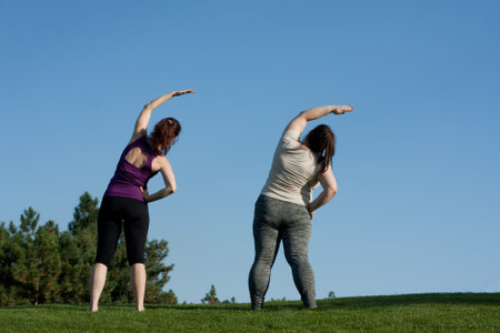 Two middle-aged women doing fitness in city park, bending body to side, blue sky background, back view. Healthy lifestyle, yoga, Pilates, weight loss. overweight woman doing yoga with trainer.の写真素材