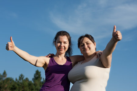 Two women hugging after good workout. Athletic woman trainer and overweight woman smiling against blue sky. Weight loss support, friendship, healthy lifestyle, outdoor sportsの写真素材