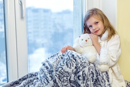 Cute little girl hugging a teddy bear. A cute baby in the room sits at the window in the winterの写真素材