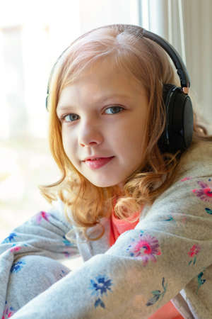 beautiful little teen girl sitting on the windowsill and listening to music on headphones with happy emotionsの写真素材
