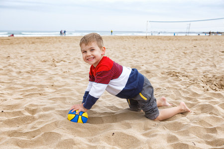 Young Boy Playing With a Colorful Beach Ball on a Sandy Shoreline During a Cloudy Afternoonの写真素材