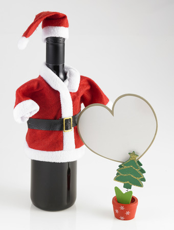 red wine bottle wearing santa claus costume with mini christmas tree holding a blank heart shape cardの写真素材