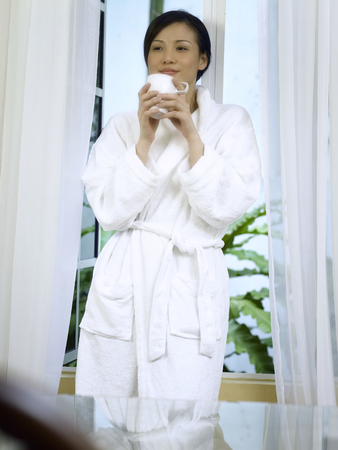 woman in bathrobe drinking coffee by a windowの写真素材