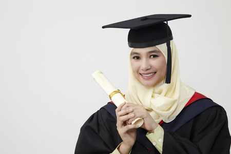 Happy young women in mortarboard holding diploma and smiling at camera while isolated on whiteの写真素材