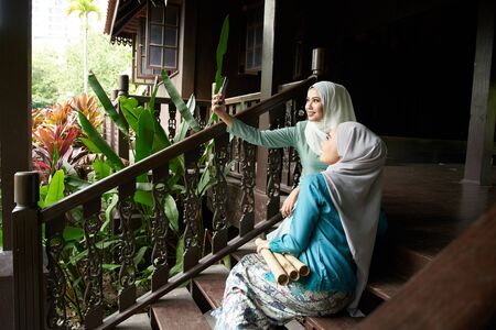 asian muslim woman wearing hijab take selfie together with smartphone at traditional malay houseの写真素材