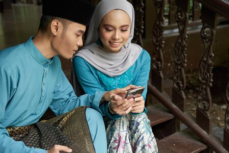 Muslim couple in traditional Malay clothing sitting at wooden stair using smart phone during Eid al-Fitr celebration.の写真素材
