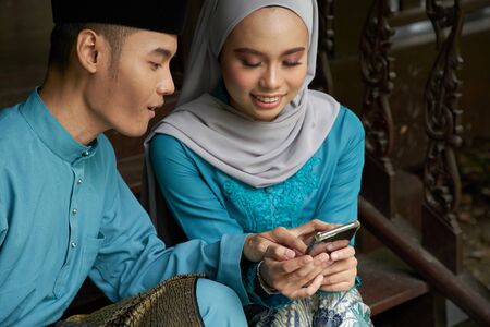 Muslim couple in traditional Malay clothing sitting at wooden stair using smart phone during Eid al-Fitr celebration.の写真素材