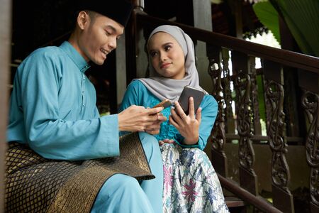 Muslim couple in traditional Malay clothing sitting at wooden stair using smart phone during Eid al-Fitr celebration.の写真素材