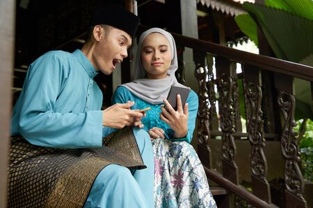 Muslim couple in traditional Malay clothing sitting at wooden stair using smart phone during Eid al-Fitr celebration.の写真素材